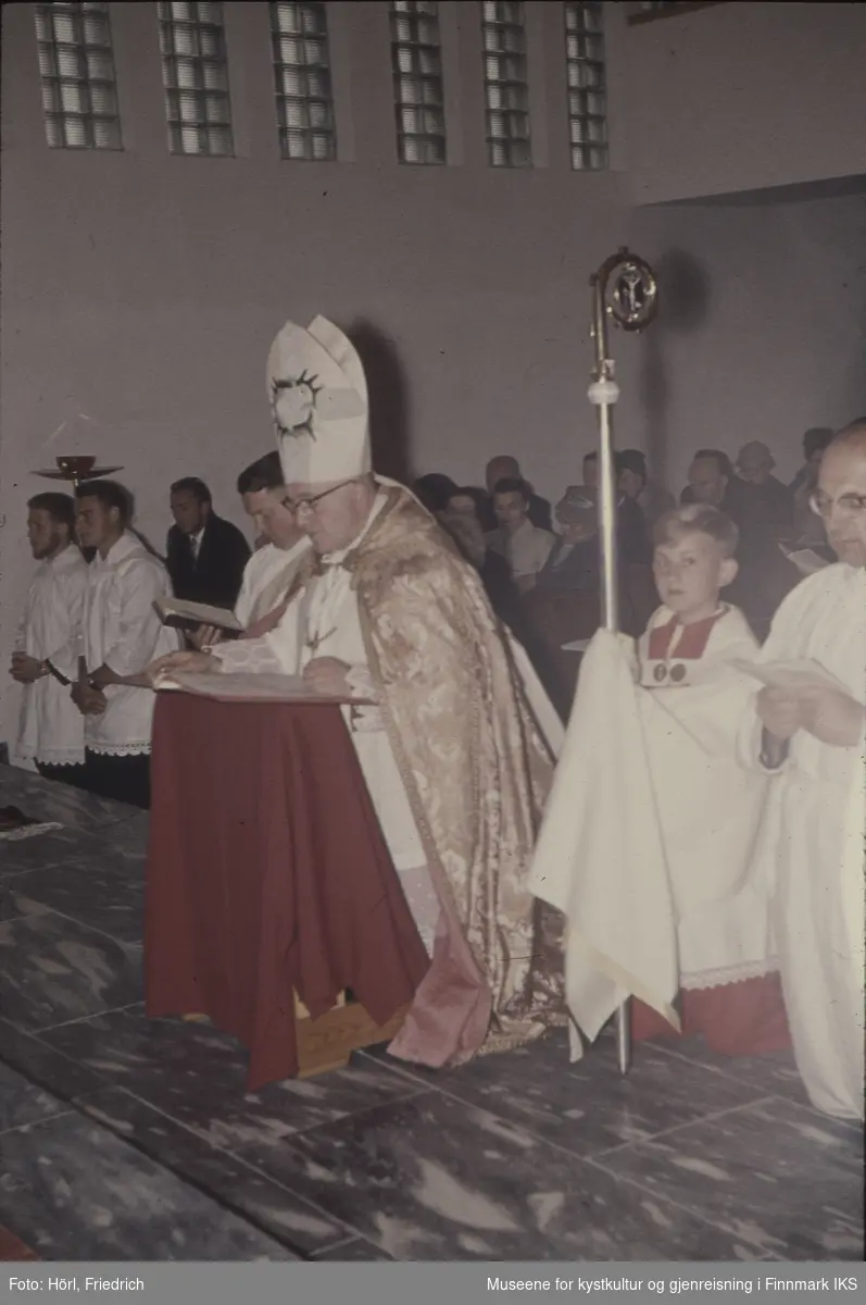 Biskop Wember kneler under bønnen ved innvielsen av Den katolske kirke St. Mikael i Hammerfest den 03.08.1958. Ved siden og bak ham kneler flere andre personer, i bakgrunnen ser man menigheten og deler av kirkerommet.