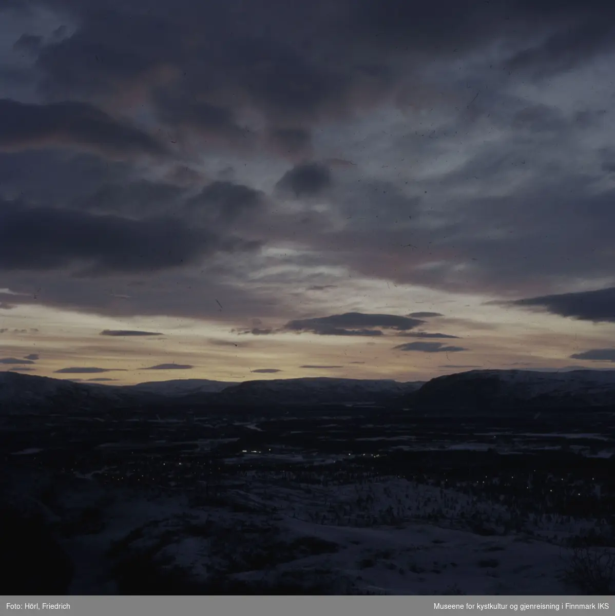 Bildet viser mest sannsynlig landskapet rundt Alta i Finnmark. Landskapet ligger snødekket i grålysningen og man ser lys i husene.  Fotografert i 1972/1973.