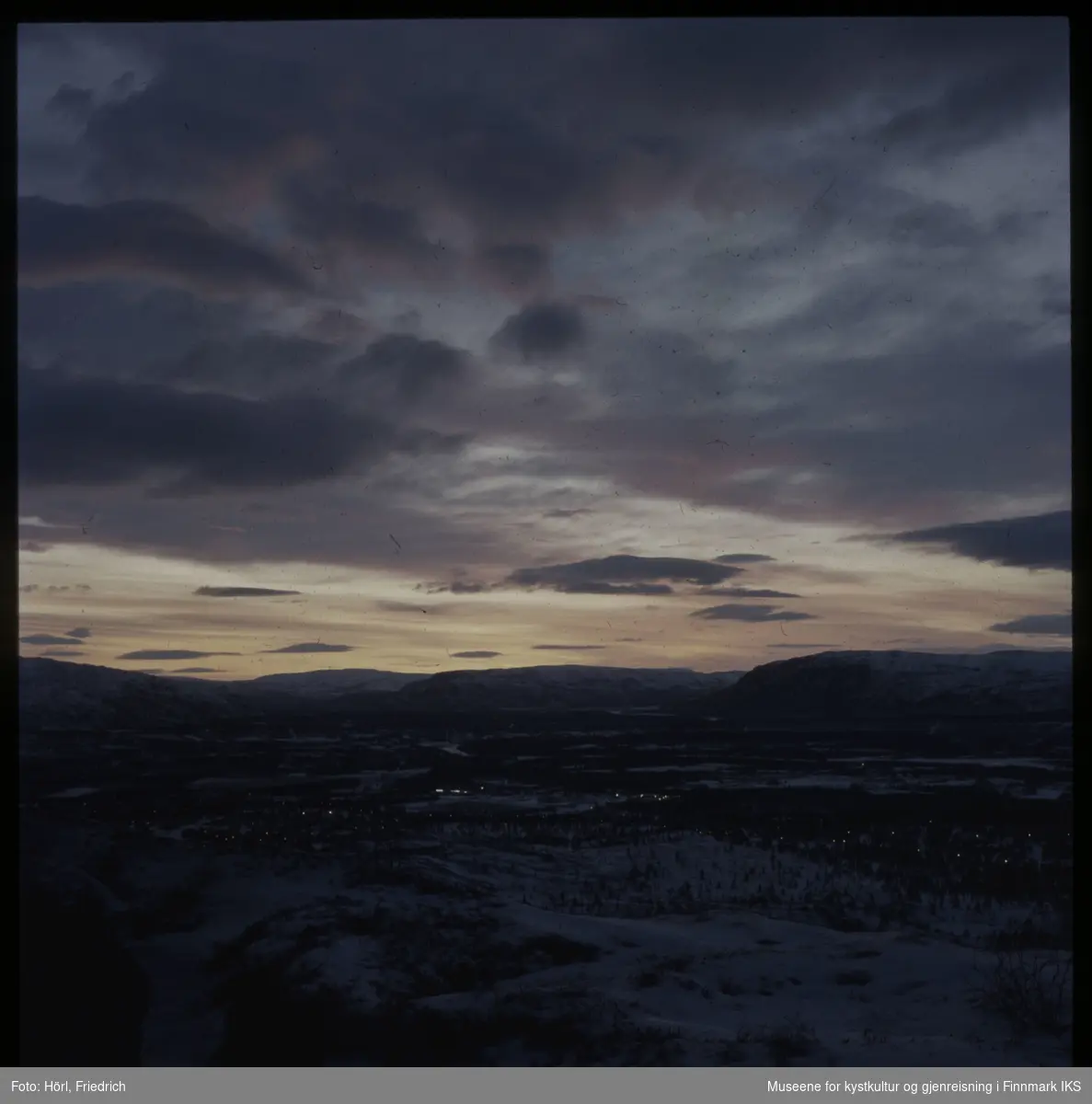 Bildet viser mest sannsynlig landskapet rundt Alta i Finnmark. Landskapet ligger snødekket i grålysningen og man ser lys i husene.  Fotografert i 1972/1973.