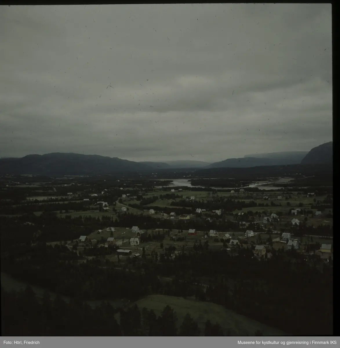 Utsikten over en dalslette med barskog, gjenreisningsbebyggelse og en elv. Mest sannsynlig er det Altaelva som er avbildet. Bildet er tatt i 1957 eller 1958.