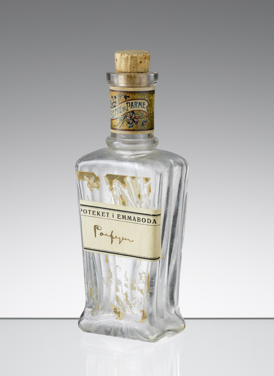 Parfymflaska med kork från Knut Lundqvists i Växjö. Stolprelief på sidorna och baksidan, samt initialerna "KL" på baksidan. Originaletiketten är bortskrapad och ersatt med en etikett från apoteket i Emmaboda, varpå man skrivit "Parfym" med bläck. Runt halsen en etikett med blommor och texten "VIOLETTE DE PARME".