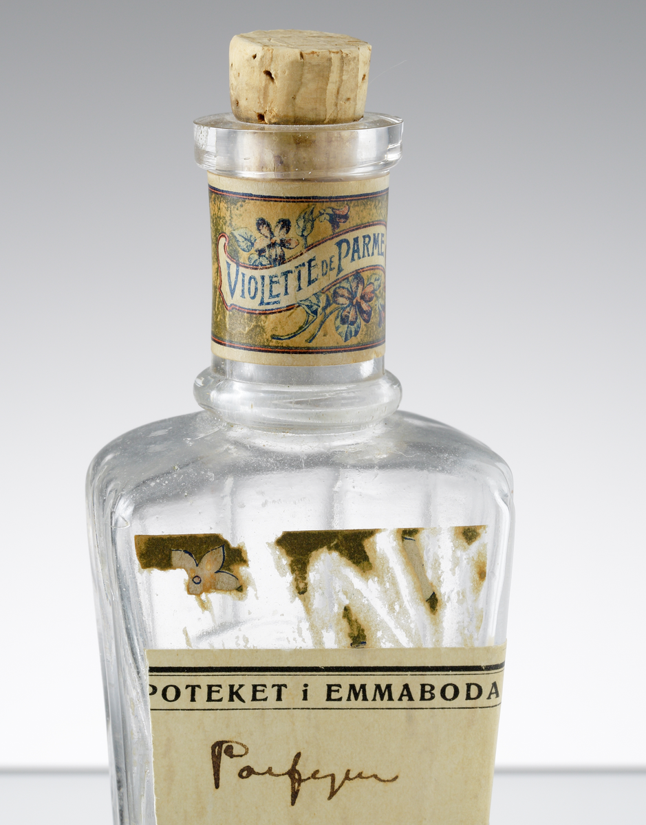 Parfymflaska med kork från Knut Lundqvists i Växjö. Stolprelief på sidorna och baksidan, samt initialerna "KL" på baksidan. Originaletiketten är bortskrapad och ersatt med en etikett från apoteket i Emmaboda, varpå man skrivit "Parfym" med bläck. Runt halsen en etikett med blommor och texten "VIOLETTE DE PARME".