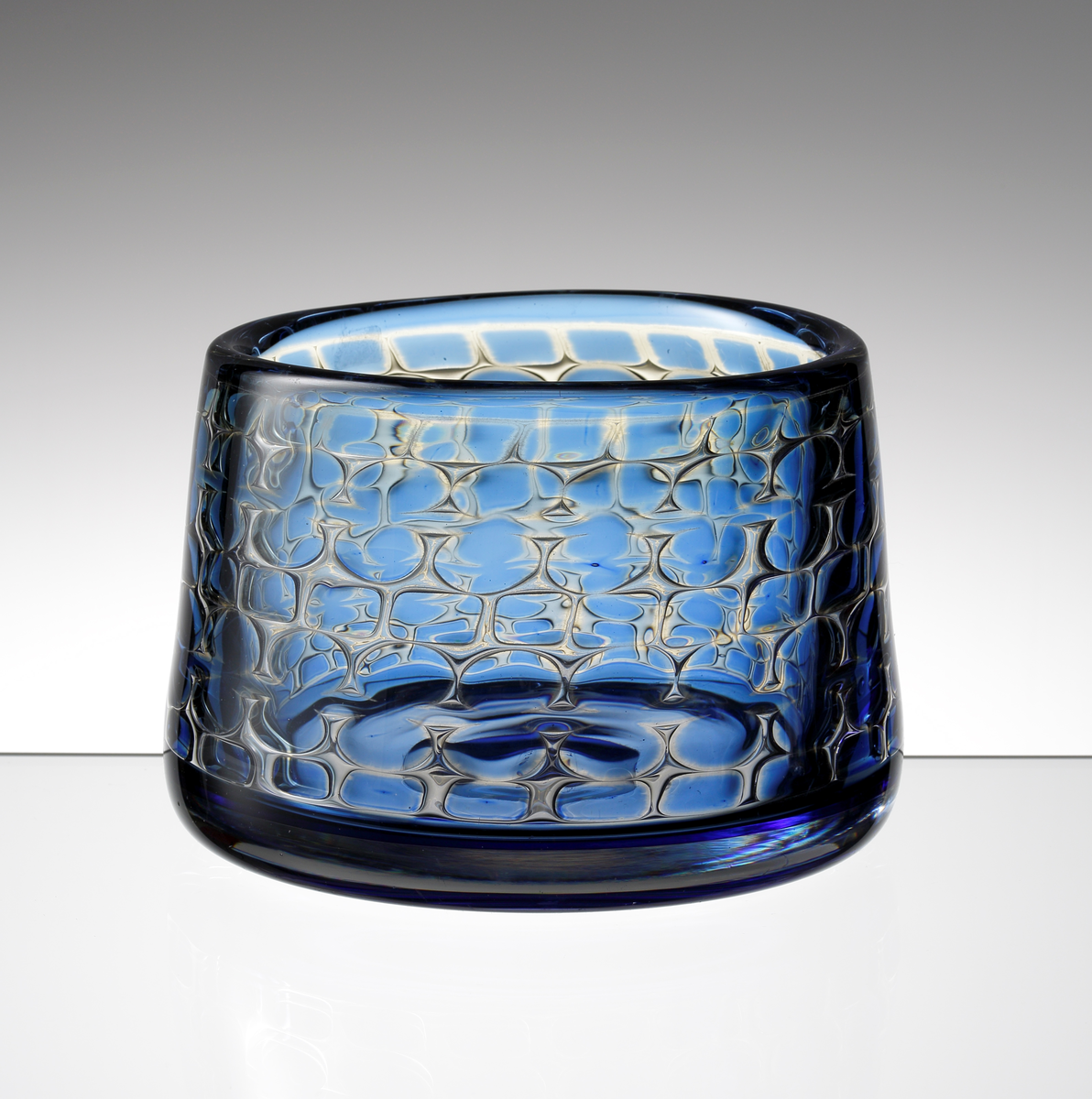 Formgiven av Ingeborg Lundin. Arielskål. Tjockväggig med cylindrisk form. Ofärgat och blått glas med geometriskt luftmönster bestående av rundlar och kvadrater.