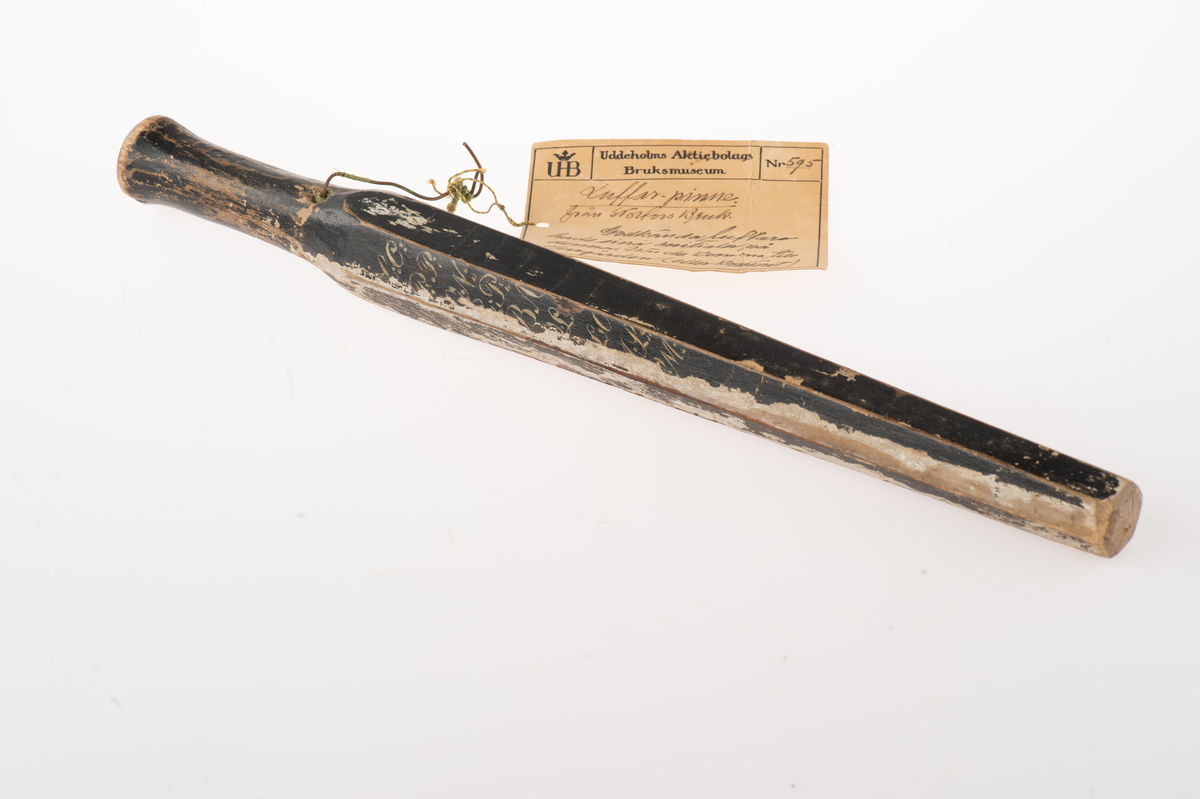 Avlångt runt svarvat trästycke med genomgående hål i ena änden. Föremålet övergår till en grövre sexkantig form som smalnar av mot ena änden. Föremålet är svartmålat med vit, endast delvis läsbar text.