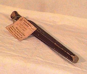 Avlångt runt svarvat trästycke med genomgående hål i ena änden. Föremålet övergår till en grövre sexkantig form som smalnar av mot ena änden. Föremålet är svartmålat med vit, endast delvis läsbar text.