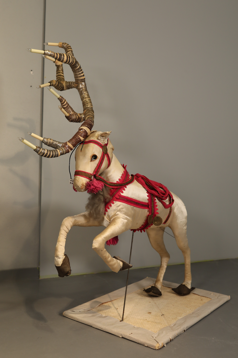 Utstillingsfigur montert på plate. Figuren er utformet som et reinsdyr i hvitt, silkeaktig stoff med brune klover og filtøyne. Geviret er surret inn med gullbånd og har sokler til lyspærer i taggene. Geviret er avtagbart. Reinsdyret har seletøy i rosa og lilla filt med gulltråd og runde metallplater. Juletrelysene til geviret er Philips 14v. Det stikker ender av ledninger ut av både kroppen og geviret. 

Reinsdyret består av flere deler:
a - kropp montert på plate med metallstøtte foran og bena skrudd fast bak. På et tidspunkt har noen stiftet fast papir over ytterkantene av det originalet stoffet.
b - gevir med plass til 8 lyspærer
c - gevir med plass til 7 lyspærer
d - løs pyntedusk
e - grå ledning
f til u - 16 lyspærer