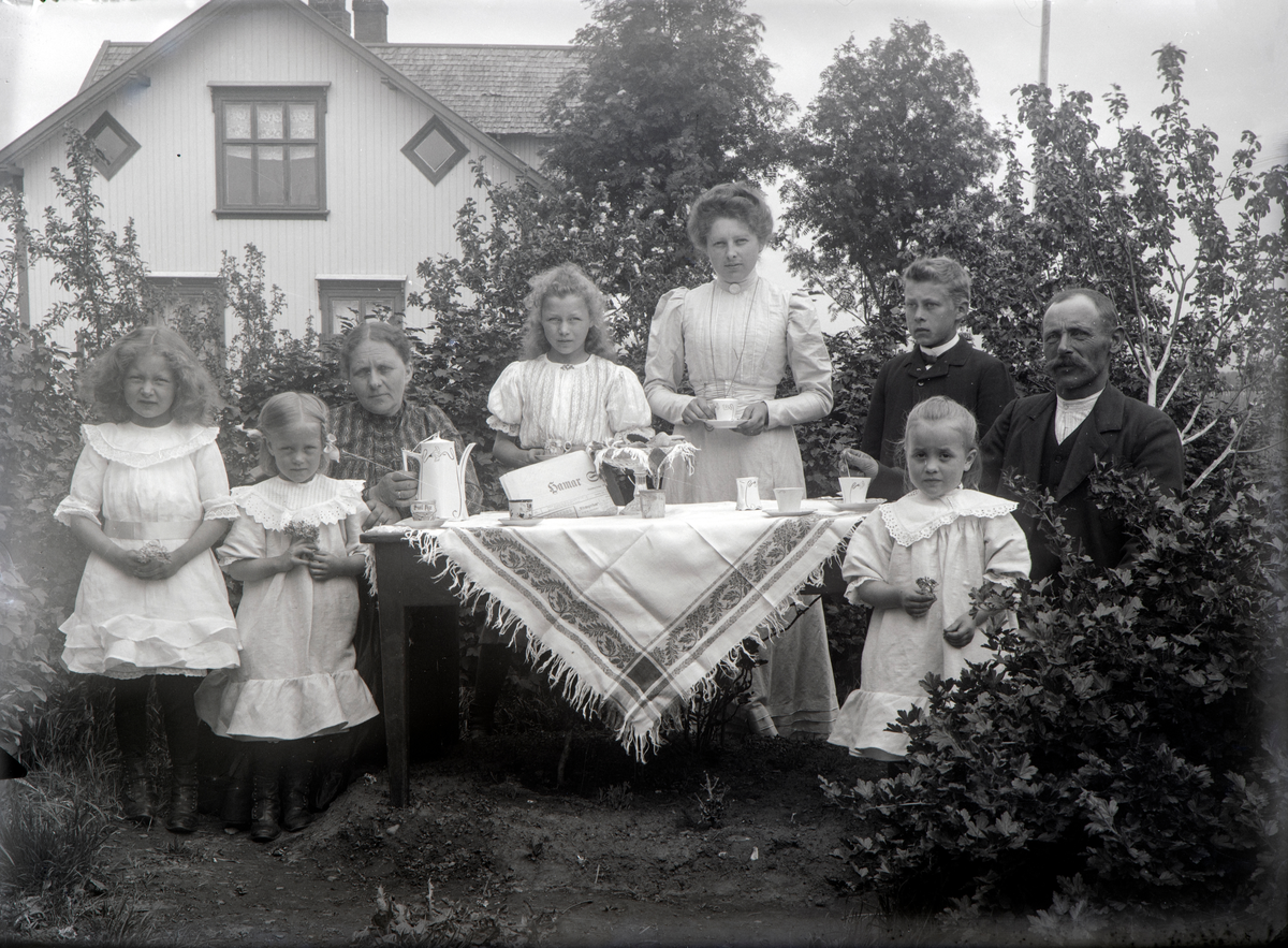 Stange, gruppe 8 kaffeselskap i hagen på Nyborg, 5 barn, byggmester Simen Nyborg (1862-1962) og Berthe Nyborg (1863-1935) og en bestemor, dekket kaffebord, Hamar Stiftstidende på bordet, barn: Oline f. 1887, Anton f. 1890, Olaf f. 1893, Oddbjørg f. 1896, Emma f. 1899, Signe Berglot f. 1901, Lilla f. 1902, 
Simen født på Lillehammer, kom til Stange 23 år gammel, tømmermester i 1887, har bygd 33  bygninger i Stangebyen, satt i Stange herredstyre fra 1890 for Arbeiderpartiet og ble æresmedlem i Stange arbeiderlag,