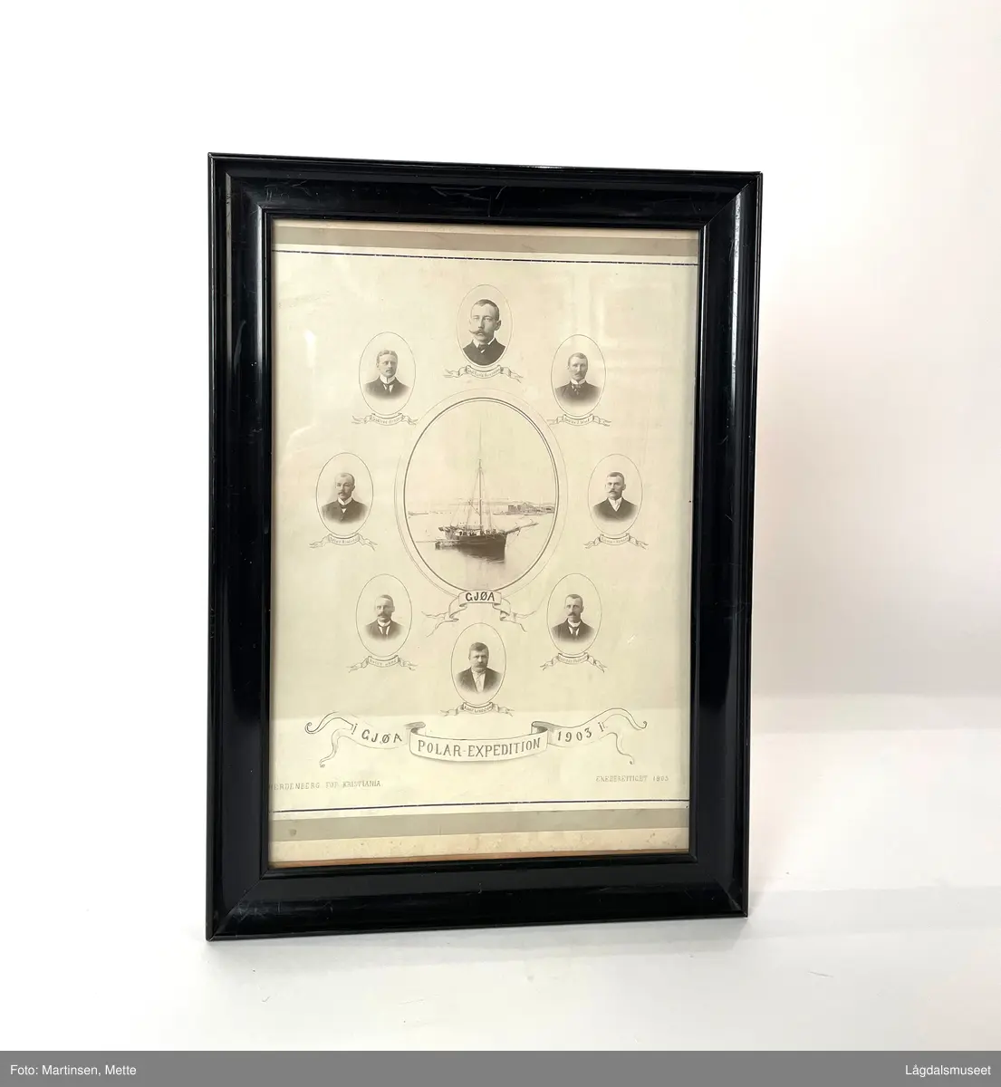 Plakat med fotografier av besetningen som var med på Gjøaekspedisjonen gjennom Nordvest-passasjen 1903-1906. Bilde av "Gjøa" i midten. Bilde av åtte menn omkranser skipet. Profilert svartlakkert ramme av tre med glass foran. Bak øyekroker og ståltråd for oppheng.