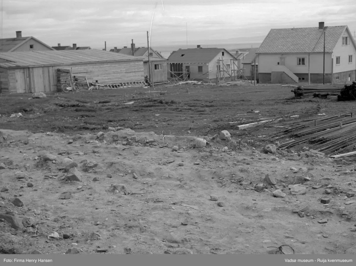 Tomta hvor Vadsø kirke skal oppføres. En del materialer og grushaug i forkant av bildet. Vi ser også gjenreisningsbrakke og gjenreisningshus. Den nye kirken vil bli oppført på samme sted som den forrige, men orientert nord-sør. Den forrige kirken ble ødelagt av krigshandlinger i oktober 1944