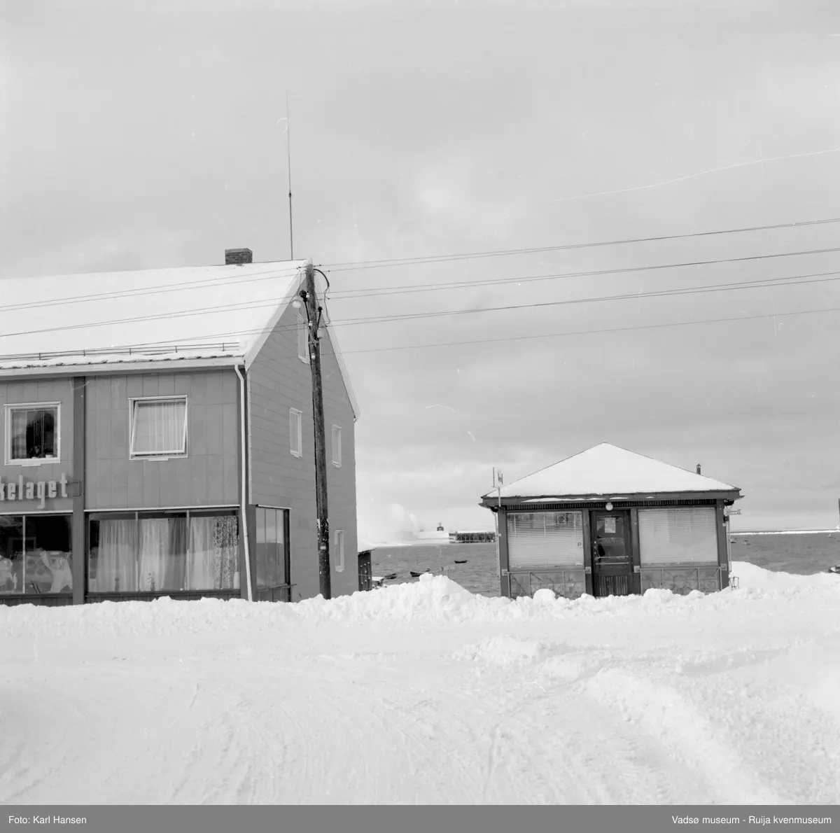 Narvesenkiosken i Tollbugata, Vadsø en vinterdag. Bildet er tatt mot vest og vi ser deler av havna m/sildoljefabrikken  og hjørnet av Samvirkelaget.