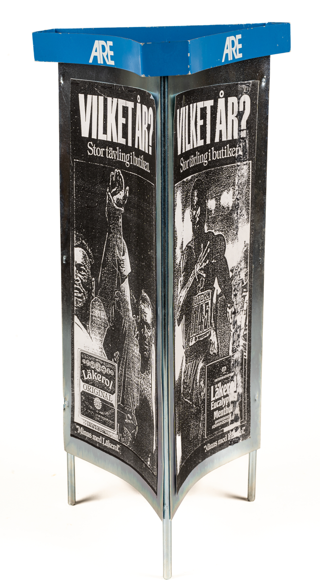 Reklamskylt, reklampelare på tre ben, i metall och papp, för Läkerol. Tre olika bilder i svart-vitt med kändisar: VILKET ÅR? Stor tävleing i butiken. 
Bilderna föreställer Ingemar Johansson "Ingo", ABBA och eventuellt Bob Hayes.