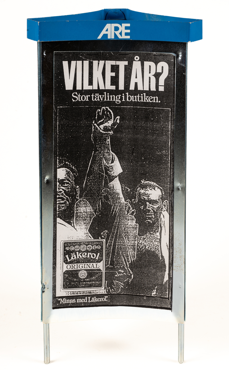 Reklamskylt, reklampelare på tre ben, i metall och papp, för Läkerol. Tre olika bilder i svart-vitt med kändisar: VILKET ÅR? Stor tävleing i butiken. 
Bilderna föreställer Ingemar Johansson "Ingo", ABBA och eventuellt Bob Hayes.