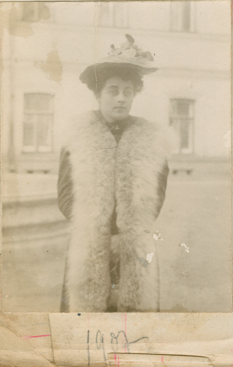 Portrett av Florentine Rostin utendørs. Hun er kledd i en lang pels-vest og hatt med blomsterdekorasjoner. I bakgrunnen er en bygning med t-post vinduer. Bildet er datert 1907.