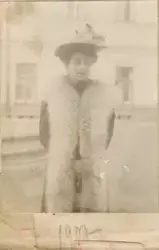 Portrett av Florentine Rostin utendørs. Hun er kledd i en lang pels-vest og hatt med blomsterdekorasjoner. I bakgrunnen er en bygning med t-post vinduer. Bildet er datert 1907.
