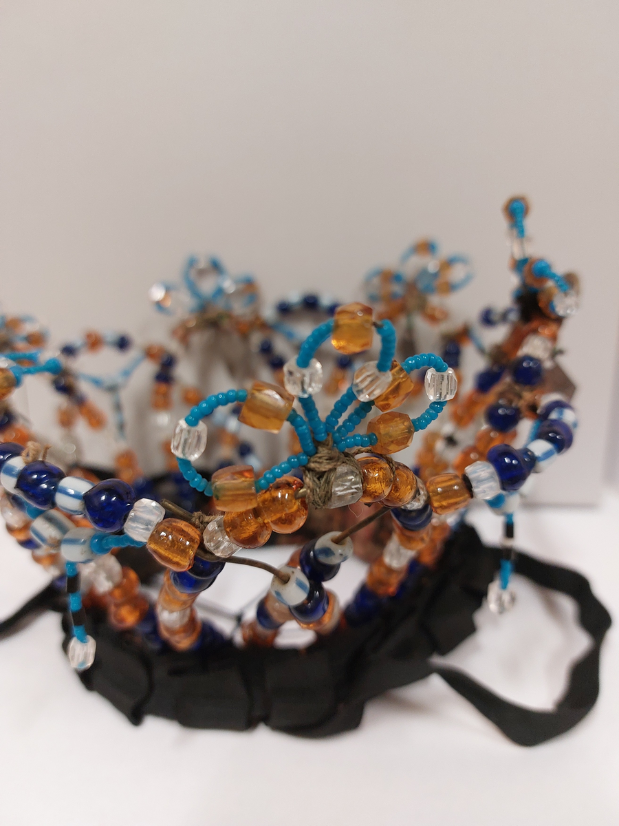 Kronen er laget av ståltråd påtredd glassperler i ulike fasonger og farger. Den består av tolv buer som står opp som går om hverandre. På toppen av annenhver bue er det laget tre mindre buer av mindre perler med innslag av større. Under et par av disse er det festet blanke knapper. Det sitter igjen heng av metall under fire av disse buene. På de andre buene er det festet heng av mindre perler med større perler på midten hvorfra det er heng av mindre perler med en større nederst.. I en av disse hengene skiller perlene seg ut fra de andre.. Krona har valk surret med, og påsydd, bånd og svart stoff. Utenpå valken er det feste et ripsbånd som er foldet til pynt. Under valken er det feste tråd i kryss. Krona har knyting i svarte ripsbånd som er sydd fast dobbelt.
