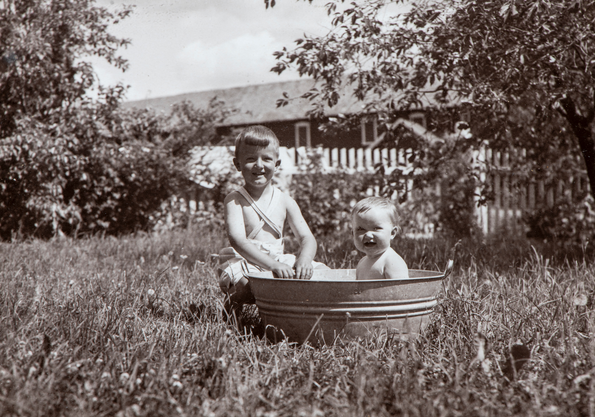 Ringsaker, Furnes, Lier gård, To barn i badebalje i hagen, fra venstre Ola Jørstad, f. 1936 og søsteren Liv Jørstad f. 1939, som bader ute. På den tida var det ikke vanlig med bad med badekar på landsbygda. Barn ble derfor badet i balje, om sommeren ofte utendørs.