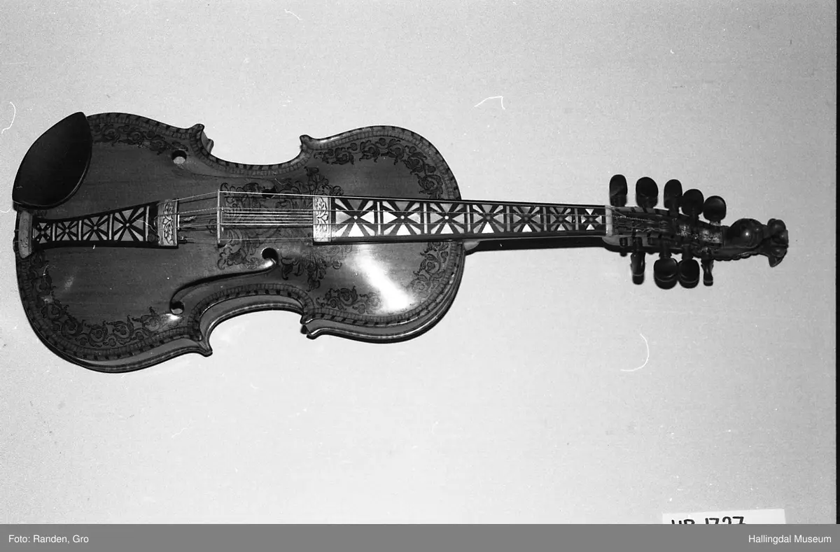 Hardingfele, tre, rødbrun lakkert. Dekorert med tusj i rosemalingsmønster, over, under, langs sidene. Gripebrett med perlemor. 7 band, 9 stemmeskruer. Halsen ender i dragehode. 5 understrenger, 3 overstrenger, samt en avslitt. Hakestøtte. Inni fela produsentens navn trykt på en papirlapp: OLAF G. HELLAND. VIOLINMAGER. NOTODDEN. Bunn påskrevet med hånd, blyant: Rep af .. R.... Kjørrdalen. BØ TELEMARK.