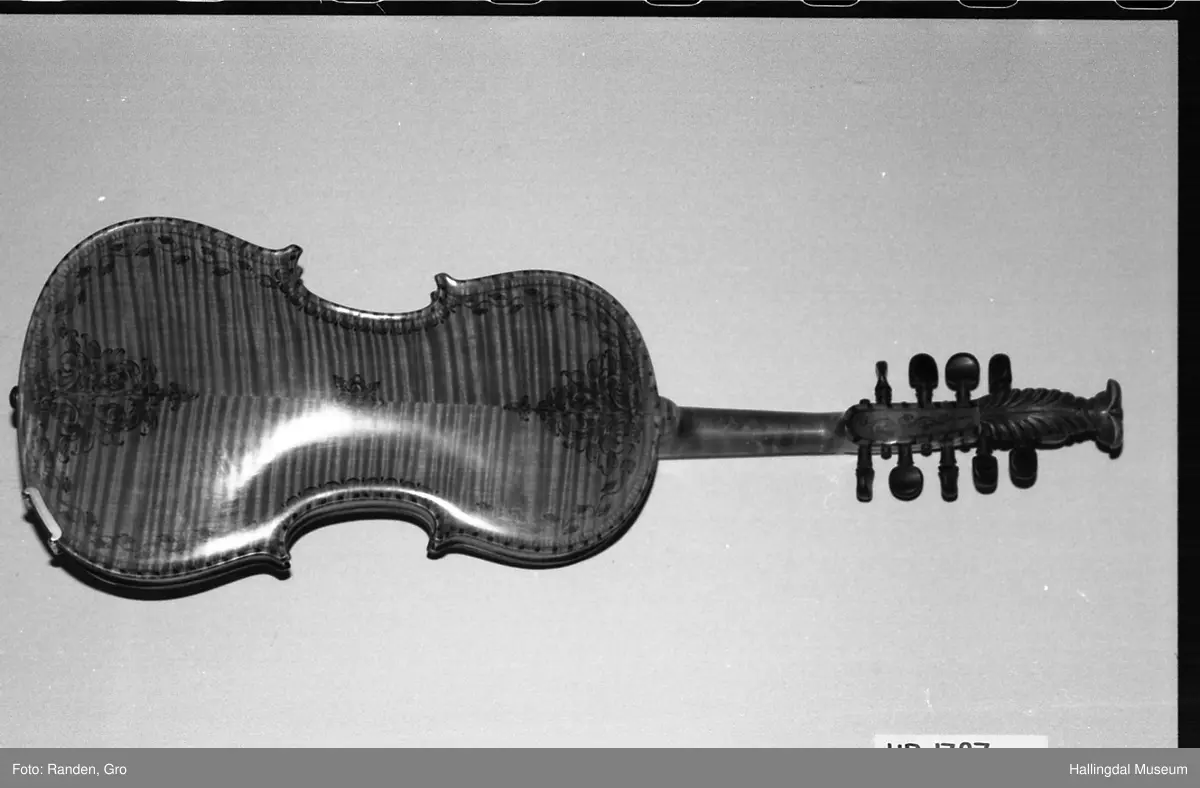 Hardingfele, tre, rødbrun lakkert. Dekorert med tusj i rosemalingsmønster, over, under, langs sidene. Gripebrett med perlemor. 7 band, 9 stemmeskruer. Halsen ender i dragehode. 5 understrenger, 3 overstrenger, samt en avslitt. Hakestøtte. Inni fela produsentens navn trykt på en papirlapp: OLAF G. HELLAND. VIOLINMAGER. NOTODDEN. Bunn påskrevet med hånd, blyant: Rep af .. R.... Kjørrdalen. BØ TELEMARK.