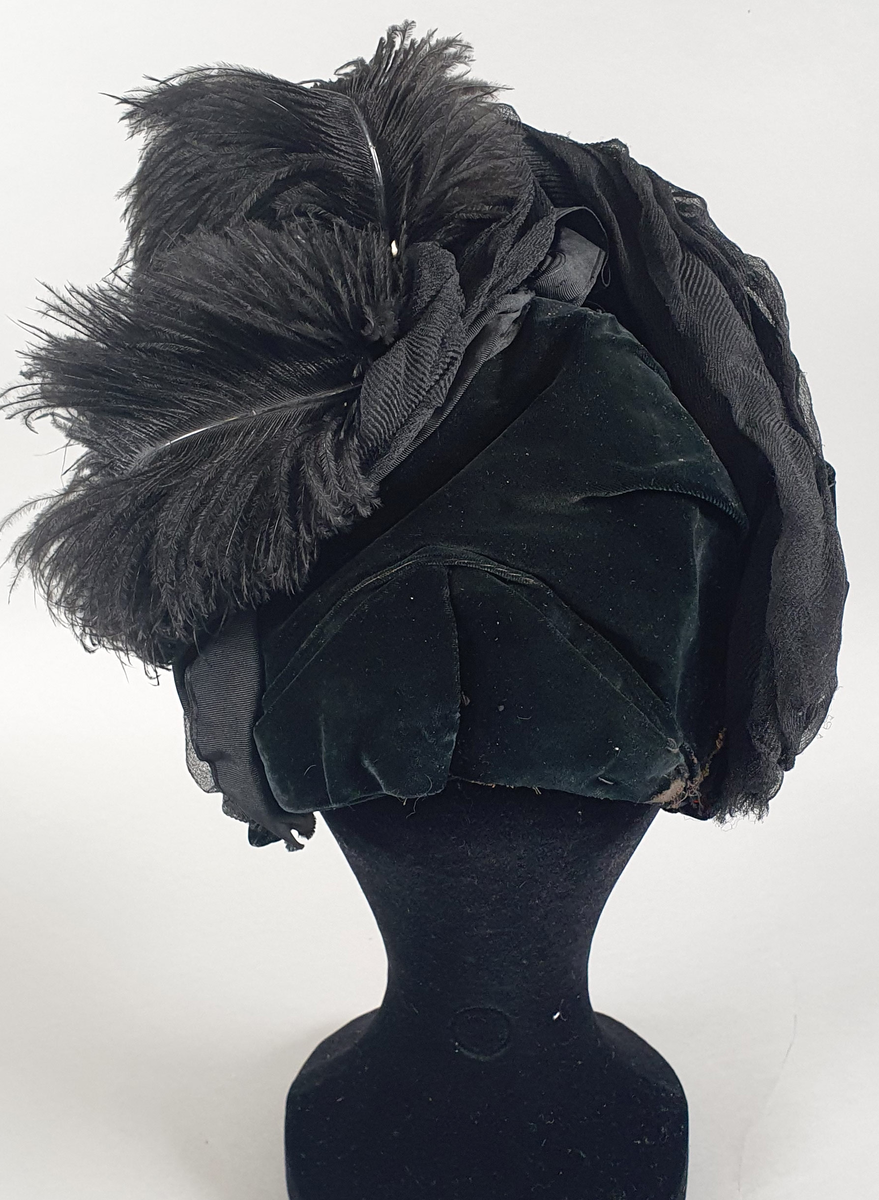 Svart damehatt av fløyel med tyll og strutsefjær, stivet opp med ståltråd. En hattenål er festet i kanten foran.