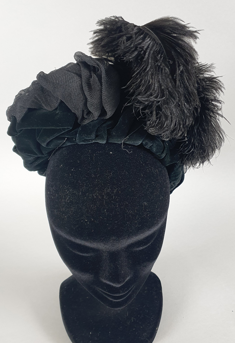 Svart damehatt av fløyel med tyll og strutsefjær, stivet opp med ståltråd. En hattenål er festet i kanten foran.