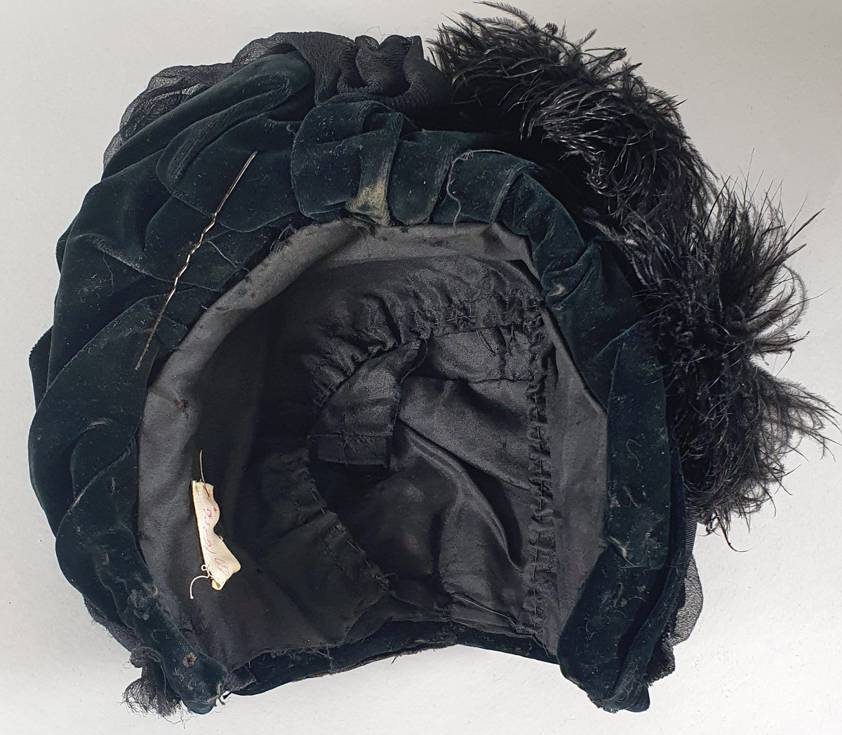 Svart damehatt av fløyel med tyll og strutsefjær, stivet opp med ståltråd. En hattenål er festet i kanten foran.