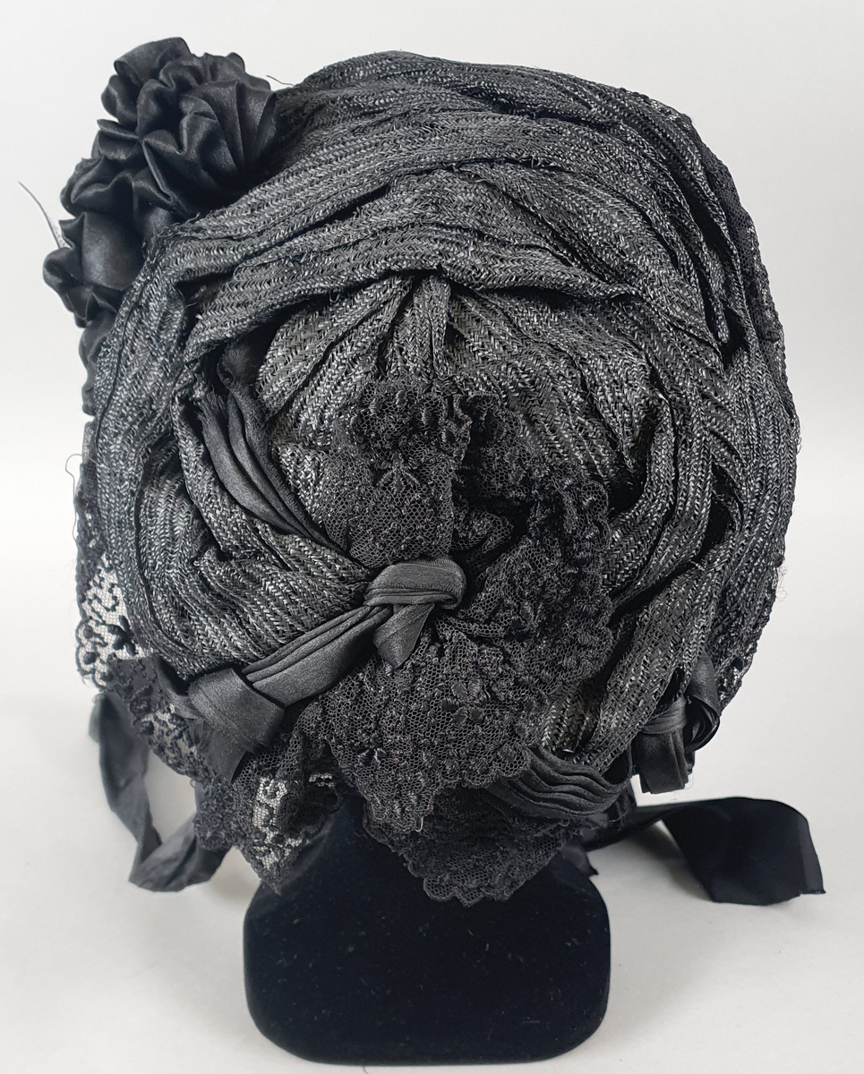 Svart damehatt av strå, silke, tyll og blonder, stivet opp med ståltråd. Lange silkesløyfer til å knyte under haken. Silkesløyfe på venstre side.