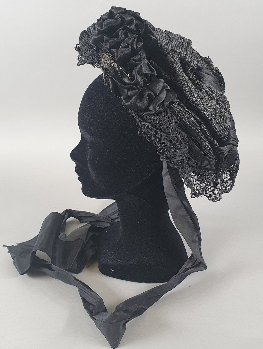 Svart damehatt av strå, silke, tyll og blonder, stivet opp med ståltråd. Lange silkesløyfer til å knyte under haken. Silkesløyfe på venstre side.