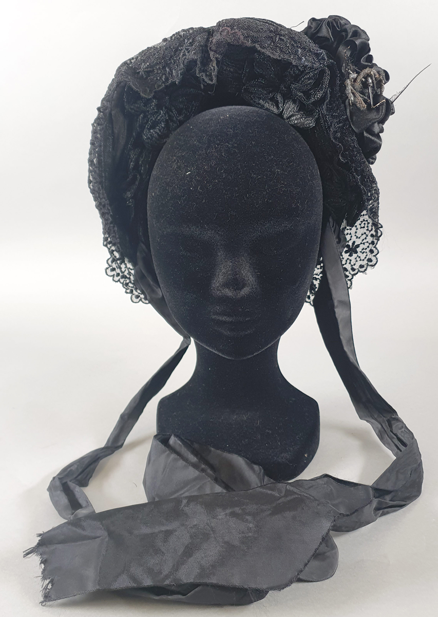 Svart damehatt av strå, silke, tyll og blonder, stivet opp med ståltråd. Lange silkesløyfer til å knyte under haken. Silkesløyfe på venstre side.