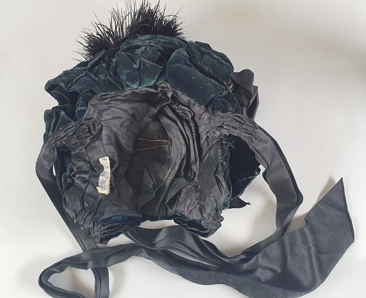 Svart damehatt av strå, silke, tyll og blonder, stivet opp med ståltråd. Lange silkesløyfer til å knyte under haken. Silkesløyfe på venstre side.