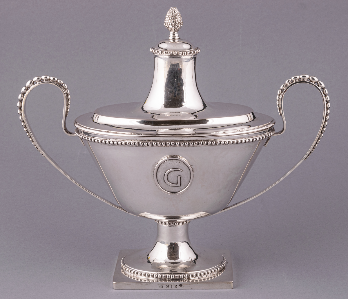 En av ett par sengustavianska sockerskålar i silver stämplade Daniel Elfbom, Gävle 1791. Oval skål på rektangulär fot. Löst lock med knopp och två utvändiga uppsvängda hänklar med pärlstav. Pålödd medaljong med Gävle stads vapen (G). Stämpar på fotens kant: DE G I 2 och tre kronor.