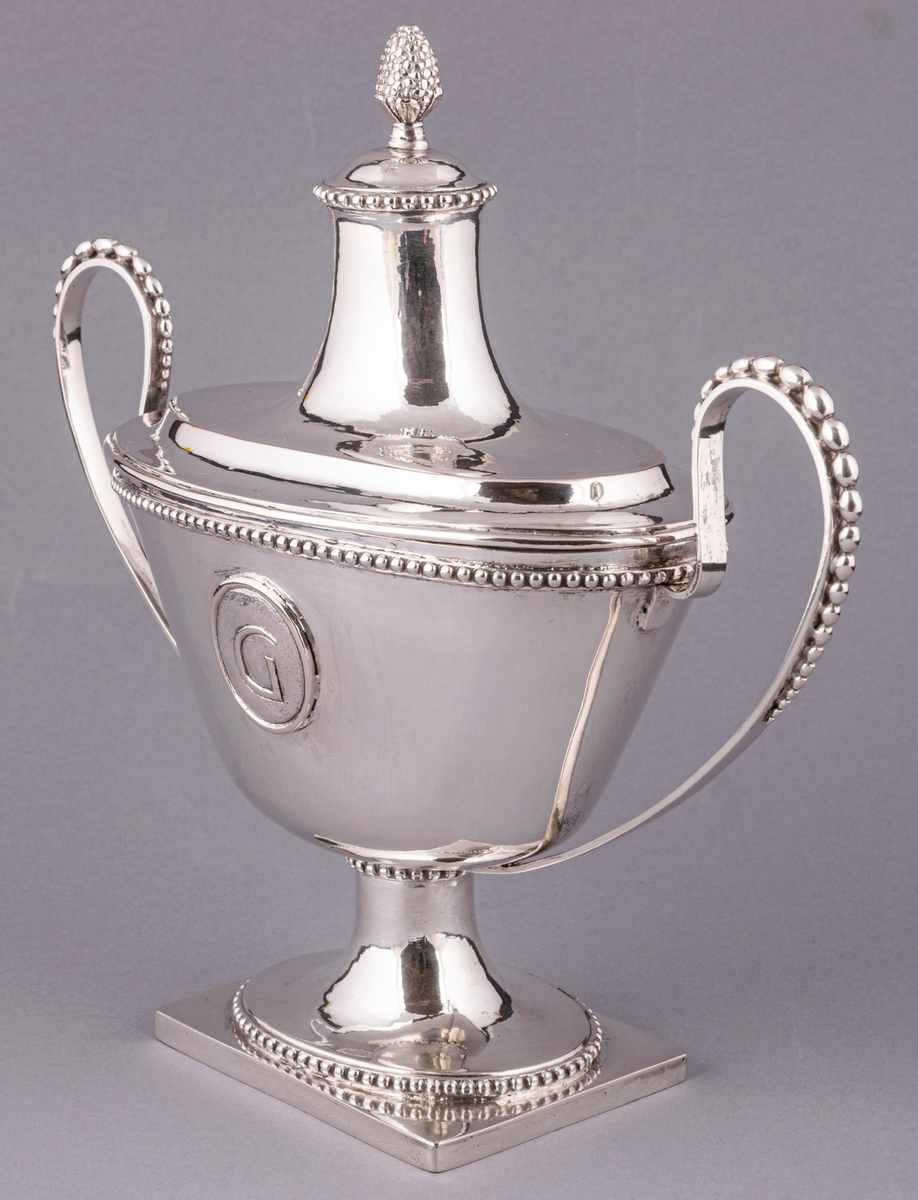 En av ett par sengustavianska sockerskålar i silver stämplade Daniel Elfbom, Gävle 1791. Oval skål på rektangulär fot. Löst lock med knopp och två utvändiga uppsvängda hänklar med pärlstav. Pålödd medaljong med Gävle stads vapen (G). Stämpar på fotens kant: DE G I 2 och tre kronor.