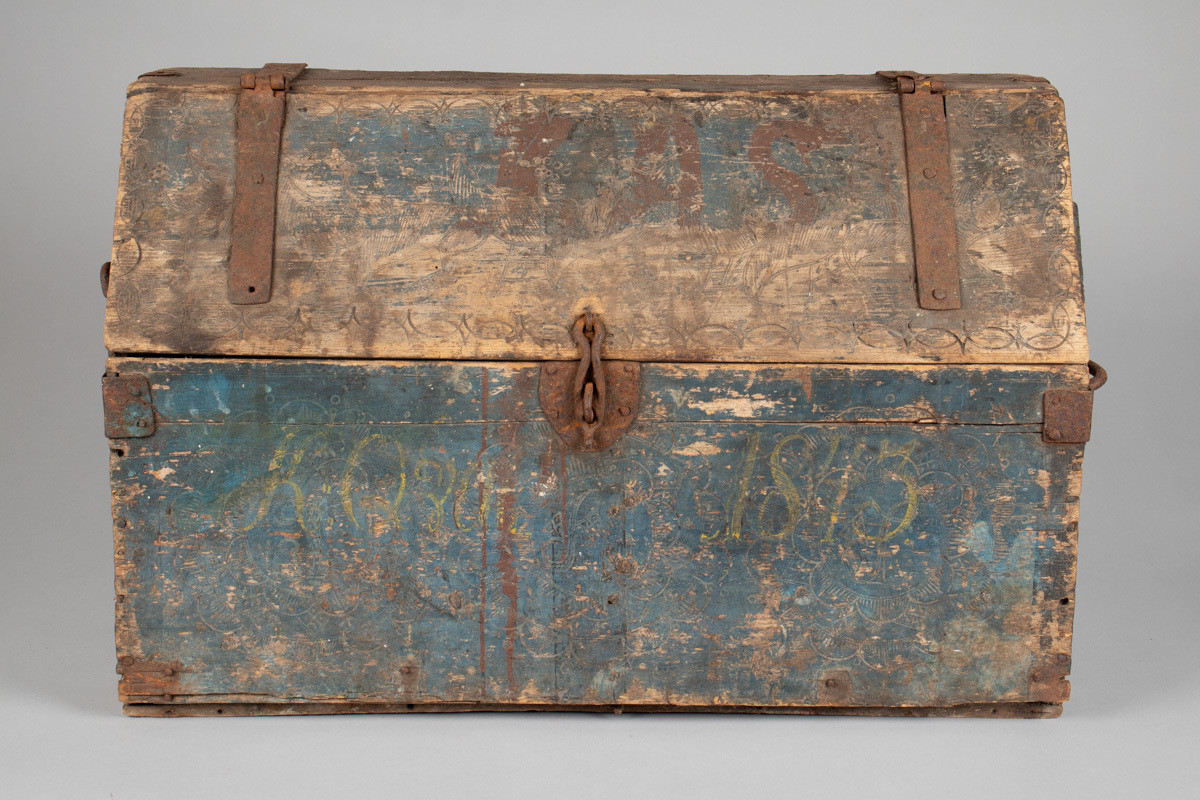 Håndhøvlet, malt og utskjært kiste i tre med jernbeslag og hektelås. Inne i kista er der tre rom, to store, og en liten skuff som har løsnet  på den ene siden.