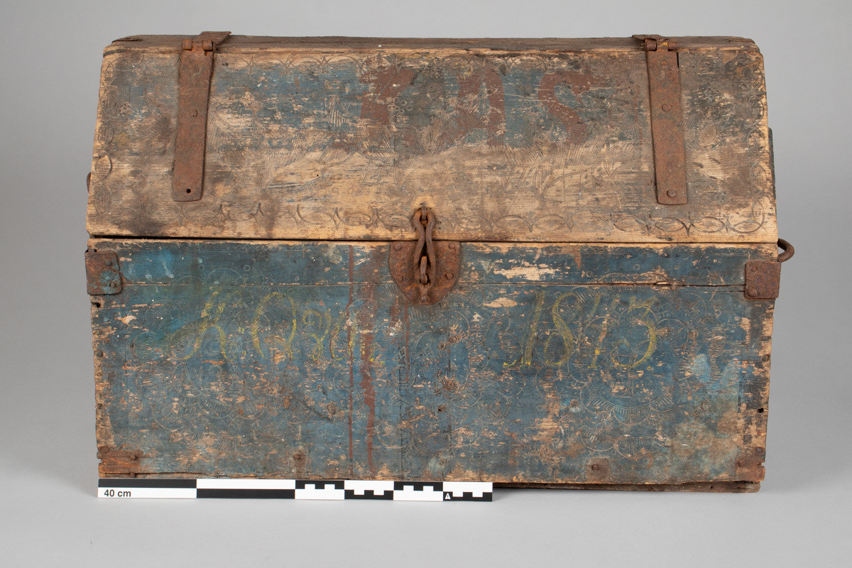 Håndhøvlet, malt og utskjært kiste i tre med jernbeslag og hektelås. Inne i kista er der tre rom, to store, og en liten skuff som har løsnet  på den ene siden.
