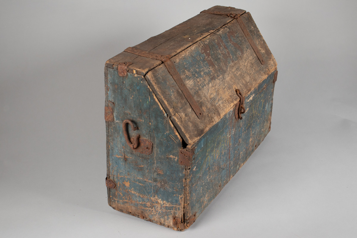 Håndhøvlet, malt og utskjært kiste i tre med jernbeslag og hektelås. Inne i kista er der tre rom, to store, og en liten skuff som har løsnet  på den ene siden.