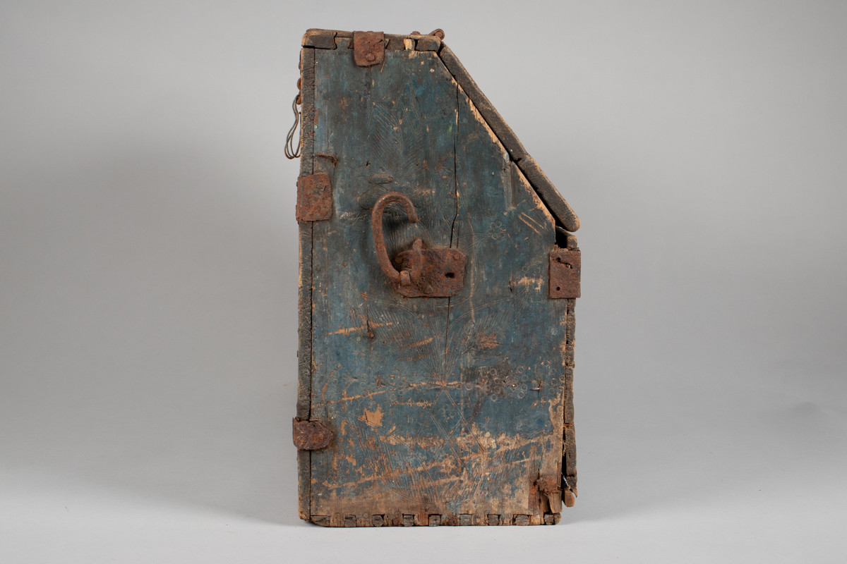 Håndhøvlet, malt og utskjært kiste i tre med jernbeslag og hektelås. Inne i kista er der tre rom, to store, og en liten skuff som har løsnet  på den ene siden.