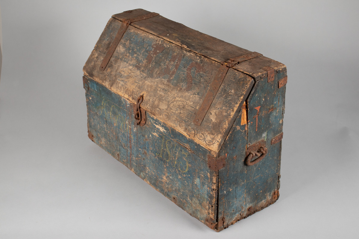 Håndhøvlet, malt og utskjært kiste i tre med jernbeslag og hektelås. Inne i kista er der tre rom, to store, og en liten skuff som har løsnet  på den ene siden.
