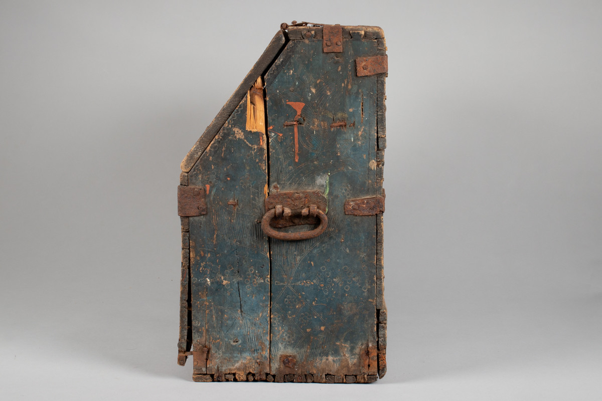 Håndhøvlet, malt og utskjært kiste i tre med jernbeslag og hektelås. Inne i kista er der tre rom, to store, og en liten skuff som har løsnet  på den ene siden.