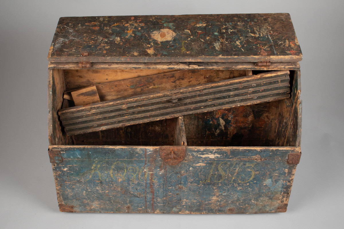 Håndhøvlet, malt og utskjært kiste i tre med jernbeslag og hektelås. Inne i kista er der tre rom, to store, og en liten skuff som har løsnet  på den ene siden.