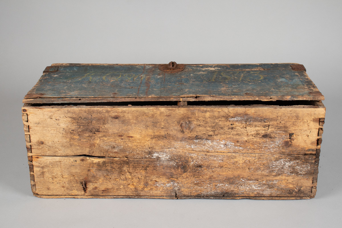 Håndhøvlet, malt og utskjært kiste i tre med jernbeslag og hektelås. Inne i kista er der tre rom, to store, og en liten skuff som har løsnet  på den ene siden.