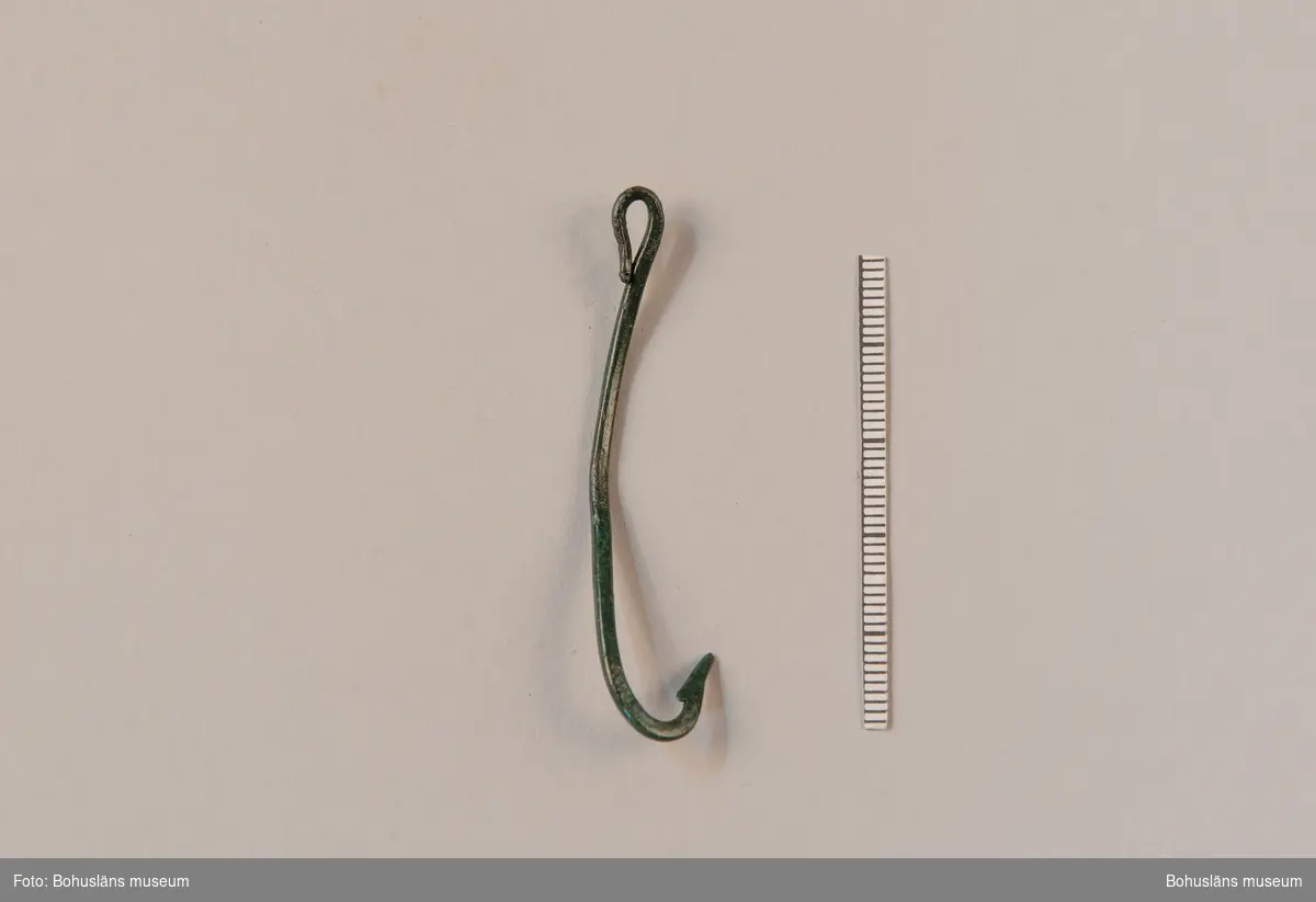 Fiskekrok av brons, tillverkad av en ten, intakt med hulling och ögla men något deformerad/böjd mitt på skaftdelen. Kroken hittades i ett utkastlager daterat till bronsålder.