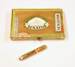 Sigarkasse. Rektangulær. Treverk-finer. Delvis dekket med papir. Oval etikett med illustrasjon av senatbygningen. Papirremse  med reklame langs sidene. Lokk innvendig med illustrasjon av mann og landskap. Mye skrift. I kassen er en sigar i selufanpapir og reklameetikett med skrift. Lokket er hengslet med tekstilbånd. Lukkes med klemme i messing.