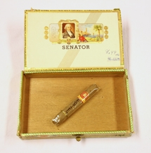 Sigarkasse. Rektangulær. Treverk-finer. Delvis dekket med papir. Oval etikett med illustrasjon av senatbygningen. Papirremse  med reklame langs sidene. Lokk innvendig med illustrasjon av mann og landskap. Mye skrift. I kassen er en sigar i selufanpapir og reklameetikett med skrift. Lokket er hengslet med tekstilbånd. Lukkes med klemme i messing.