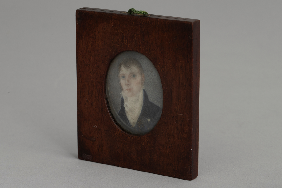 Firkantet ramme av antagelig mahogny med oval åpning på begge sider. På den ene side under glass et mannlig brystbilde en face i empiredrakt, ifølge katalogen malt på elfenben. På den andre side under glass et empiremotiv: urne på sokkel i landskap laget i perlemor med små perler, metalldeler osv. på blå bakgrunn. En hempe av flettet grønn tråd er limt i overkant mellom de to rammedelene.