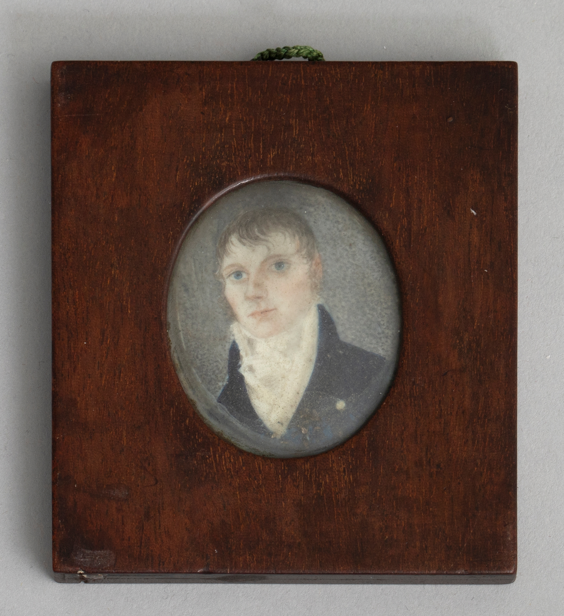 Firkantet ramme av antagelig mahogny med oval åpning på begge sider. På den ene side under glass et mannlig brystbilde en face i empiredrakt, ifølge katalogen malt på elfenben. På den andre side under glass et empiremotiv: urne på sokkel i landskap laget i perlemor med små perler, metalldeler osv. på blå bakgrunn. En hempe av flettet grønn tråd er limt i overkant mellom de to rammedelene.