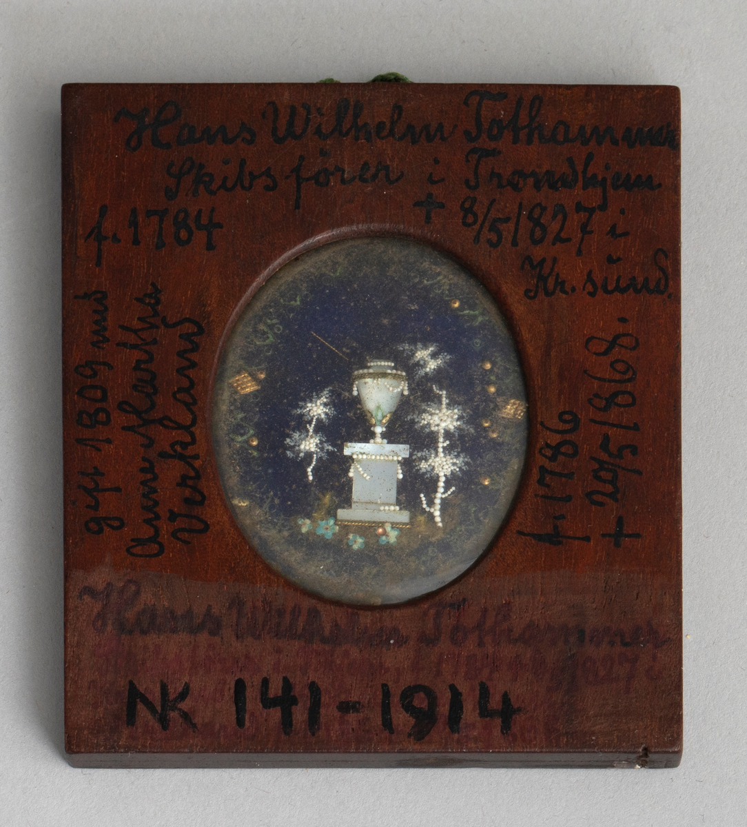 Firkantet ramme av antagelig mahogny med oval åpning på begge sider. På den ene side under glass et mannlig brystbilde en face i empiredrakt, ifølge katalogen malt på elfenben. På den andre side under glass et empiremotiv: urne på sokkel i landskap laget i perlemor med små perler, metalldeler osv. på blå bakgrunn. En hempe av flettet grønn tråd er limt i overkant mellom de to rammedelene.