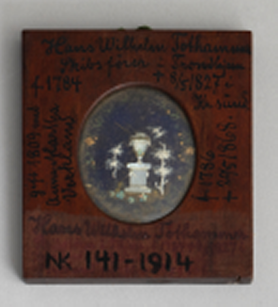 Firkantet ramme av antagelig mahogny med oval åpning på begge sider. På den ene side under glass et mannlig brystbilde en face i empiredrakt, ifølge katalogen malt på elfenben. På den andre side under glass et empiremotiv: urne på sokkel i landskap laget i perlemor med små perler, metalldeler osv. på blå bakgrunn. En hempe av flettet grønn tråd er limt i overkant mellom de to rammedelene.