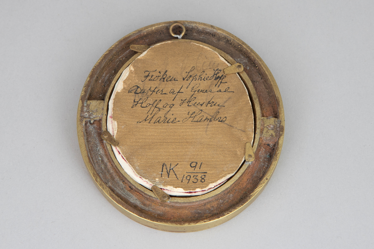 Kvinneportrett i oval ramme av bronse; brystbilde i halv profil mot høyre. Portrettet er uført i oljemaling på skinn. Kvinnen har på seg en svart utringet kjole med perlebroderi, rosa rose og lyst sjal ved venstre skulder. Hun bærer et svart bånd med perler og stener rundt halsen. Maleriet er under glass, rammen er profilert, med en skråriflet kant ved innerste profil. Forgylt papplate med påskrift på baksiden.
Tilhørende eske med hengslet lokk. Esken er trukket med rødt skinn, innvendig rød silke og fløyel. Innvendig i lokket forgylt trykk: "Sophie Hoff, datter av General Nikolai Hoff og hustru Marie Hambro".