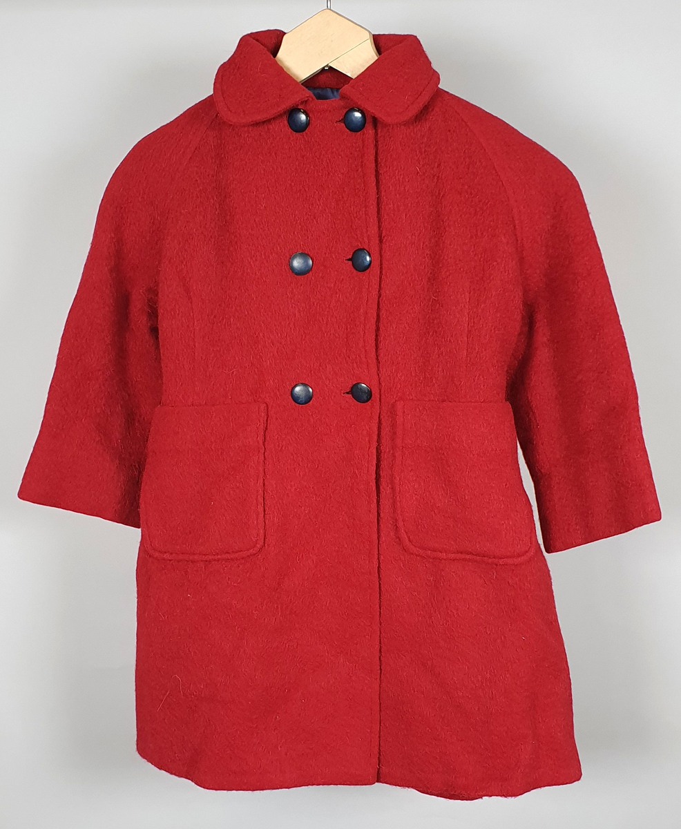 Rød kåpe av ullfilt for jente. Dobbeltspent med svarte knapper, påsydde lommer, raglanermer og skjortekrage. Fôret med blå kunstsilke. Til kåpen følger en hatt der pullen er satt sammen av seks trekanter, og med brem med syv parallelle sømmer.