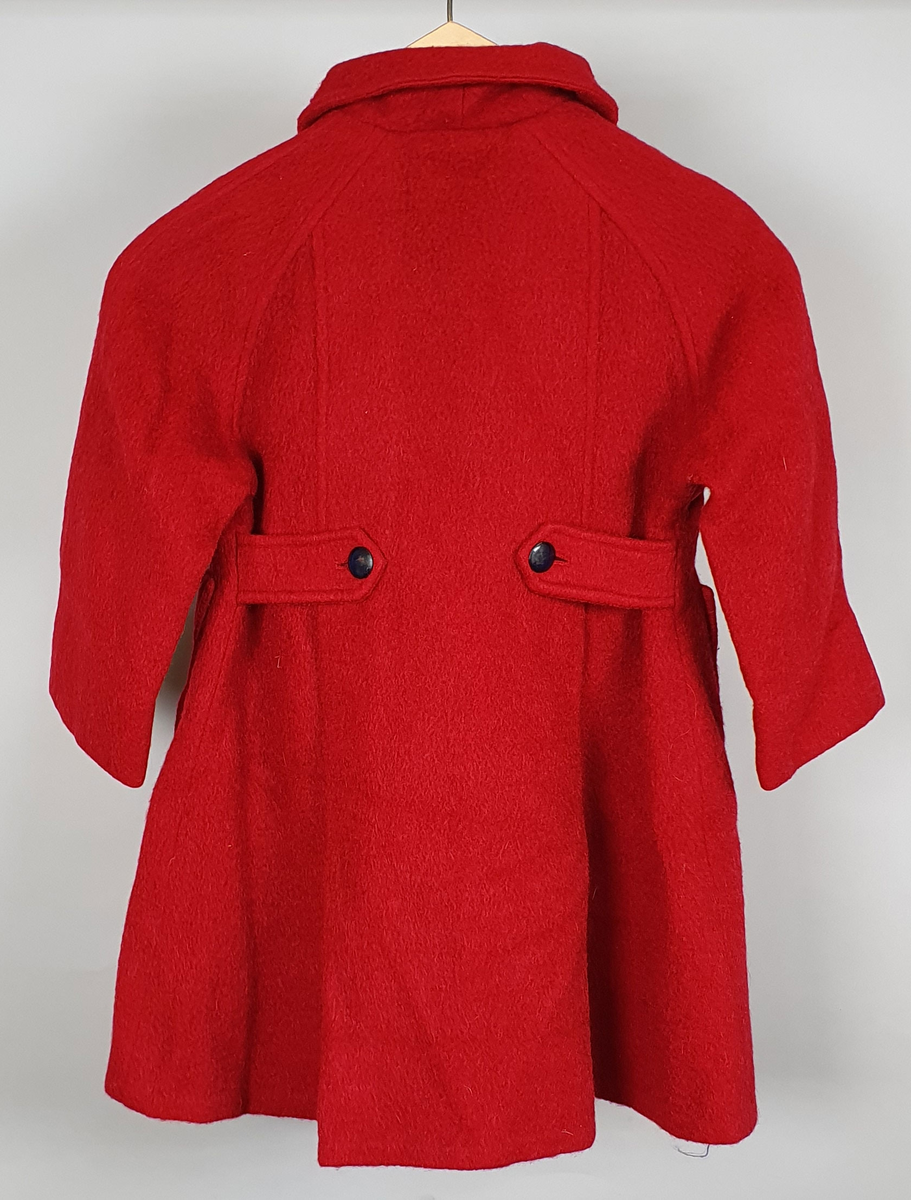 Rød kåpe av ullfilt for jente. Dobbeltspent med svarte knapper, påsydde lommer, raglanermer og skjortekrage. Fôret med blå kunstsilke. Til kåpen følger en hatt der pullen er satt sammen av seks trekanter, og med brem med syv parallelle sømmer.