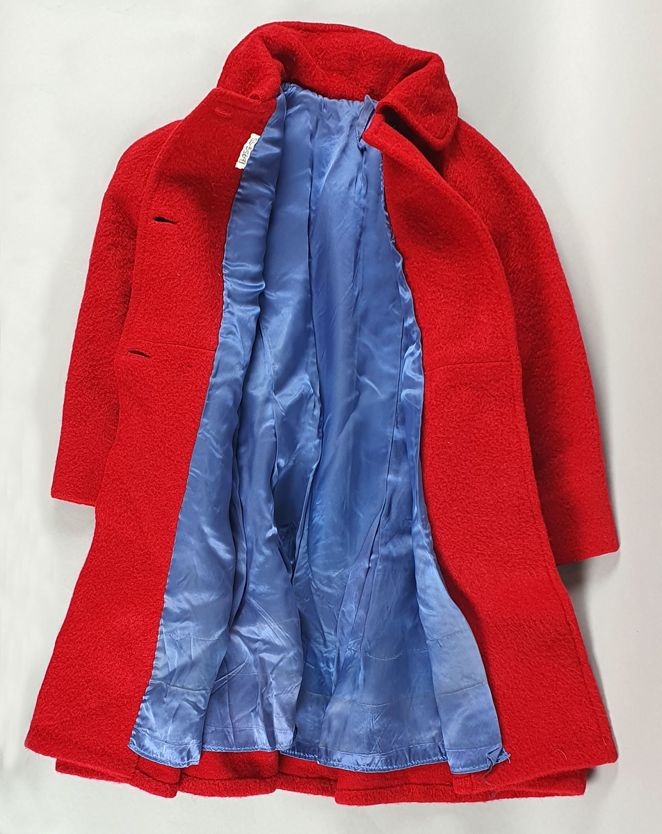 Rød kåpe av ullfilt for jente. Dobbeltspent med svarte knapper, påsydde lommer, raglanermer og skjortekrage. Fôret med blå kunstsilke. Til kåpen følger en hatt der pullen er satt sammen av seks trekanter, og med brem med syv parallelle sømmer.